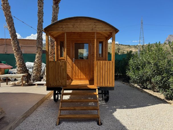 Entrada frontal del Circus Wagon 5 m Caravana de madera con escaleras, rodeada de palmeras y vegetación. Cielo azul despejado.