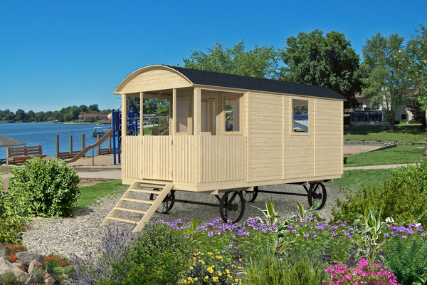 Circus Wagon instalado en entorno glamping Caravana de madera beige con ruedas, ubicada junto a un lago y rodeada de flores.