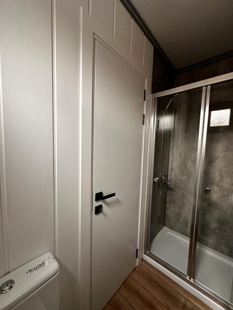 Puerta de baño junto a una ducha con mampara de cristal y un suelo de madera.